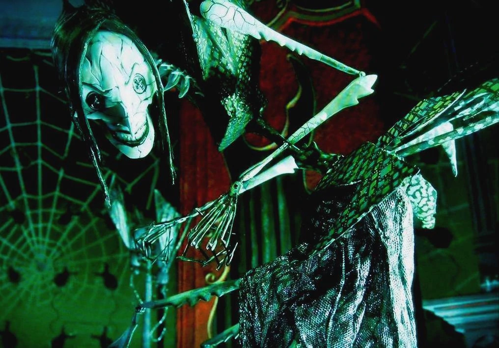 The Other Mother from Coraline, with inhumanly long insectoid arms & legs, hands made of needles, cracked-pottery skin and buttons for eyes. She is leaning menacingly towards the camera.